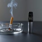 “A lit cigarette in an ashtray next to a pod-style vape device on a tabletop under natural light.”